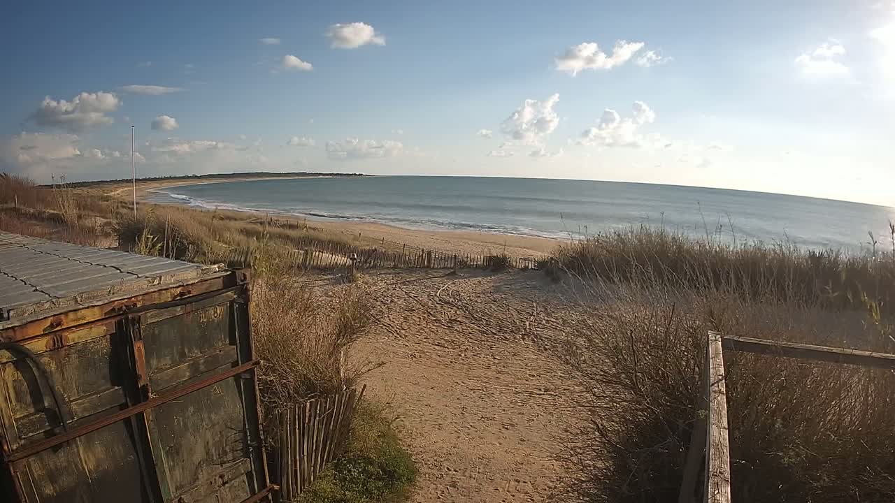 A sandy beach path leads to a calm ocean under a partly cloudy sky, with a weathered metal container visible in the foreground.