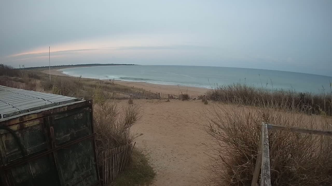 A sandy beach with calm, teal water stretches along a coastline under an overcast sky, viewed from behind a weathered container and a wooden fence.
