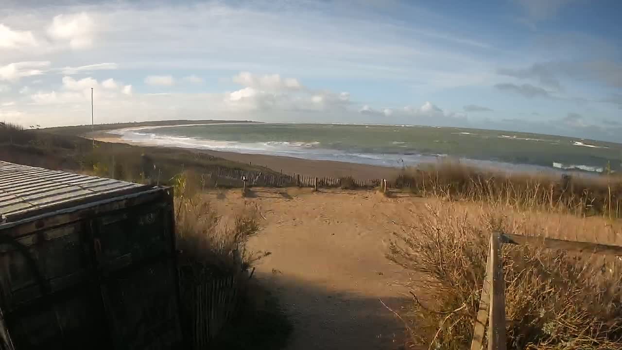 A view from behind a dark-colored shipping container shows a sandy path leading to a beach with choppy waves under a partly cloudy sky.