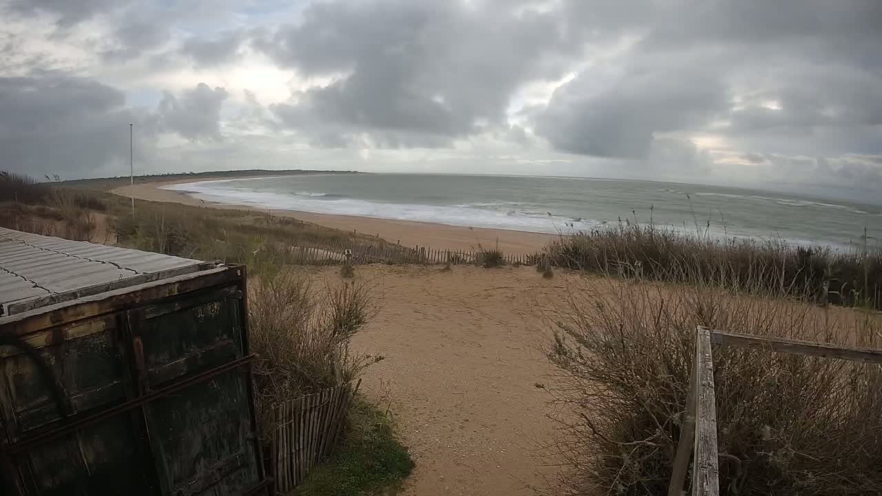 A view from a dune shows a wavy ocean under a cloudy sky, with a sandy beach and sparse vegetation in the foreground, partially obscured by a weathered container.