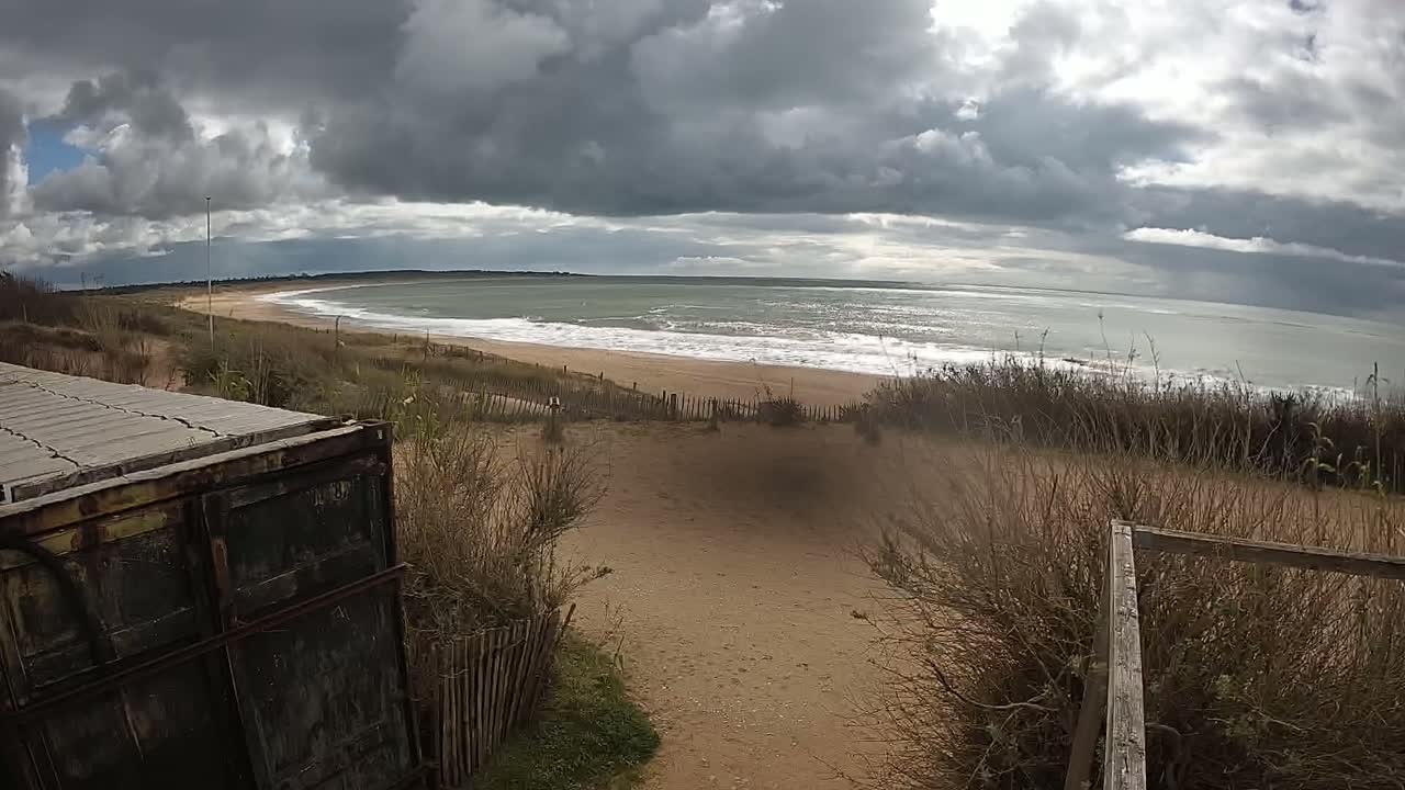 A sandy beach with gentle waves under a cloudy sky is viewed from behind a weathered container and wooden fence.