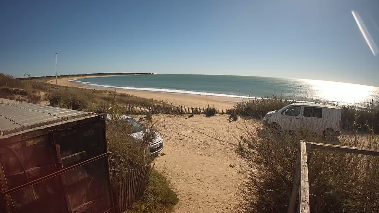 A sandy beach with gentle waves under a clear blue sky is viewed from behind a rusty container, with two parked vehicles nearby.
