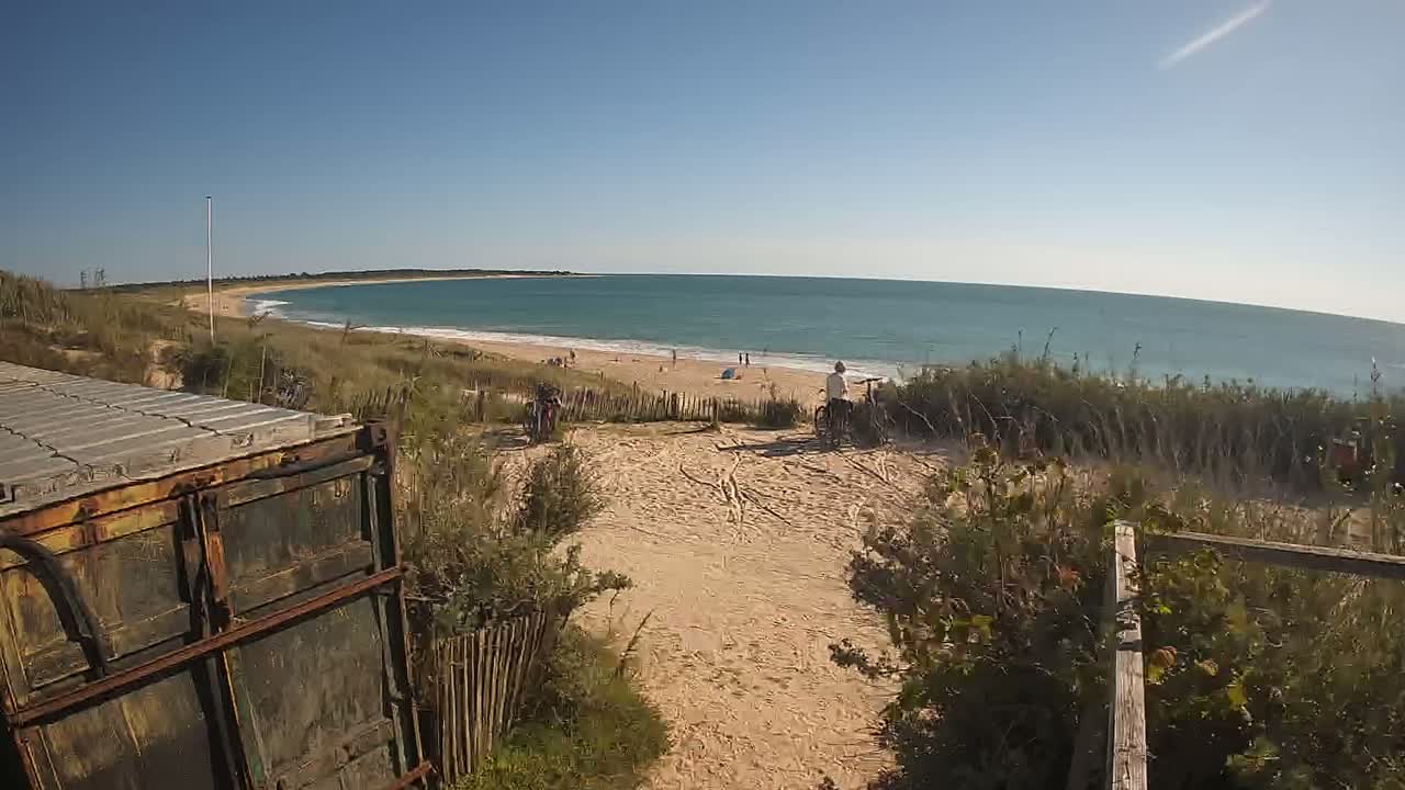 A sandy path leads to a calm ocean beach on a sunny day, with a few people visible on the shore and some bicycles parked nearby.