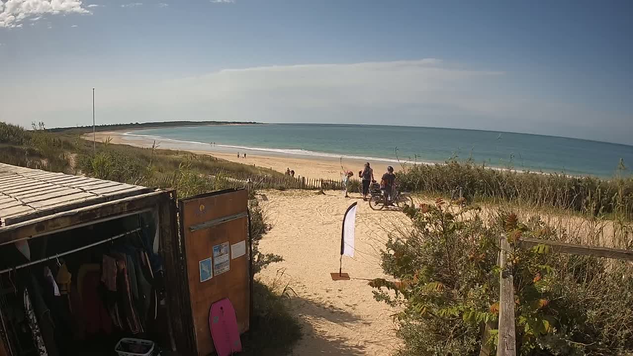 A sandy beach path leads to a calm ocean under a partly cloudy sky, with people near bicycles and a shipping container used as a changing room nearby.