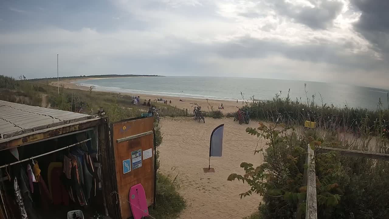 A sandy beach with people and several bicycles is visible beyond a repurposed shipping container used for storage, under a cloudy sky.