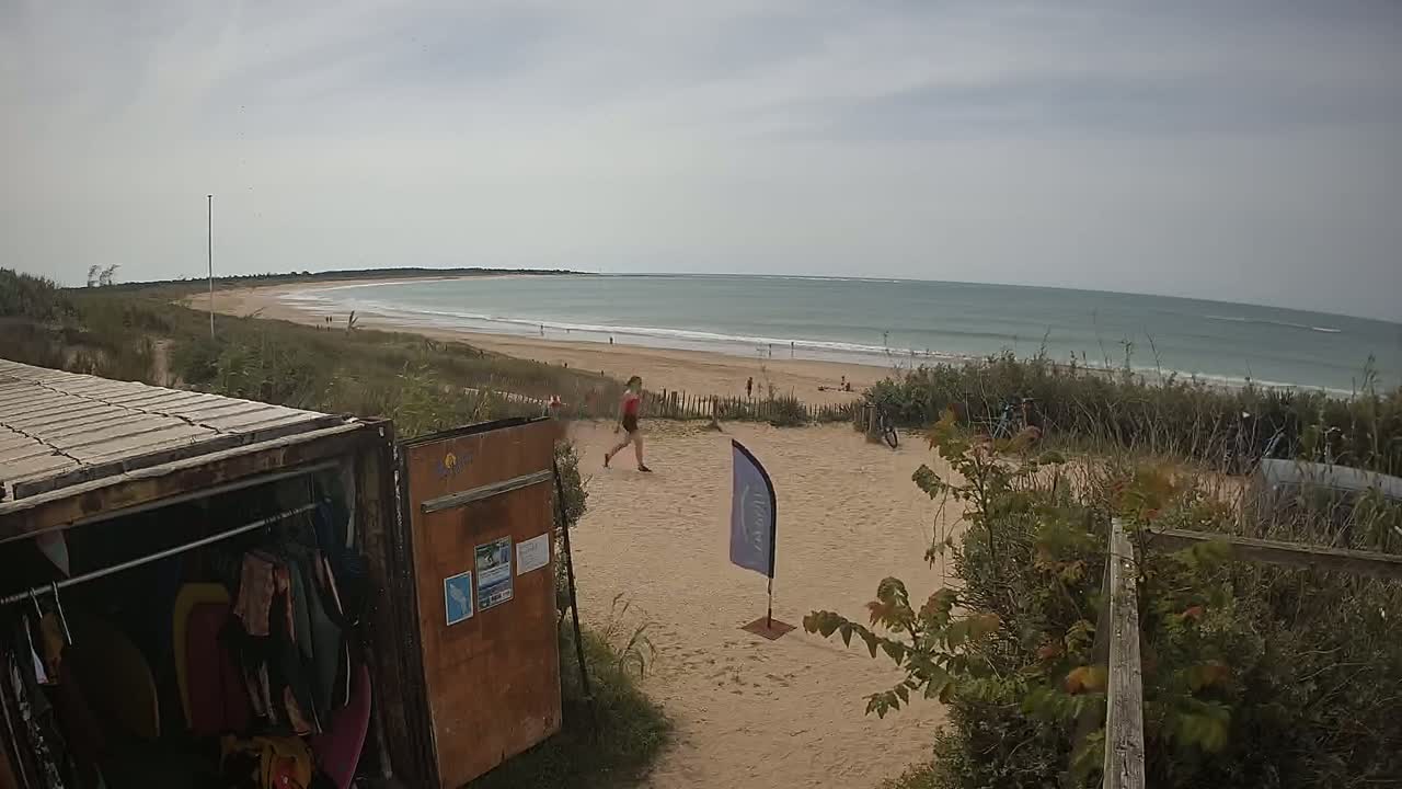 A woman walks on a sandy area near a beach with calm ocean waves under an overcast sky; a storage container with clothing is visible in the foreground.