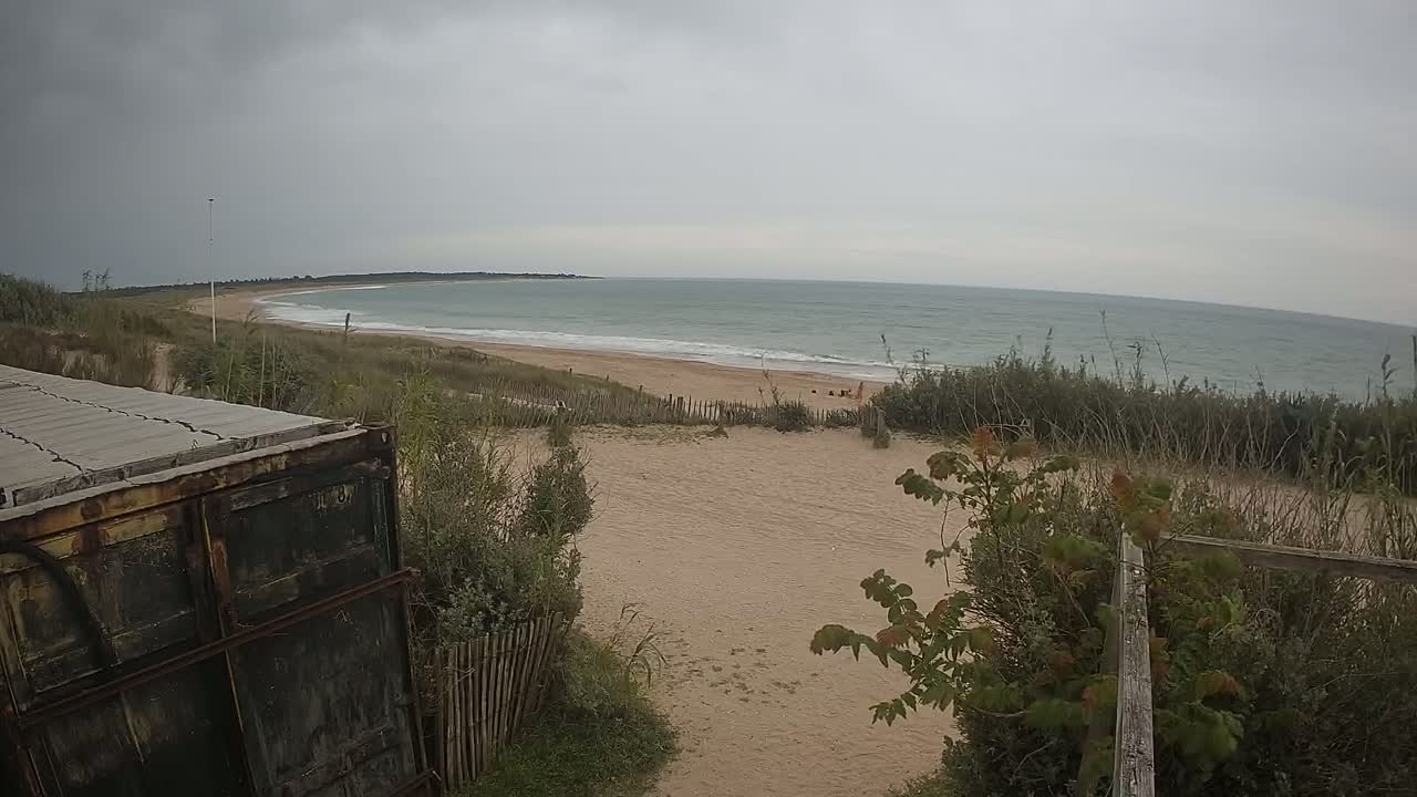 A sandy beach with a calm sea under an overcast sky is viewed from behind a weathered container and a wooden fence.