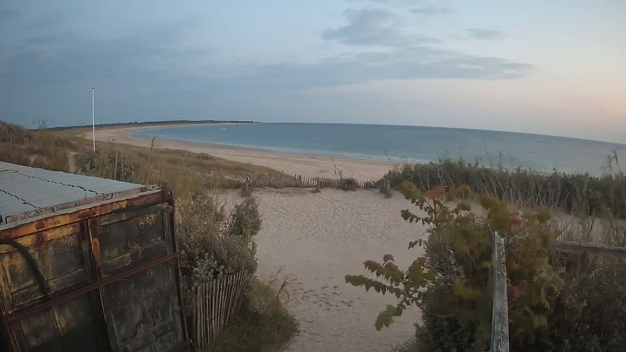 A sandy beach with calm ocean waters under a mostly cloudy sky is viewed from behind a rusty metal container and a wooden fence.