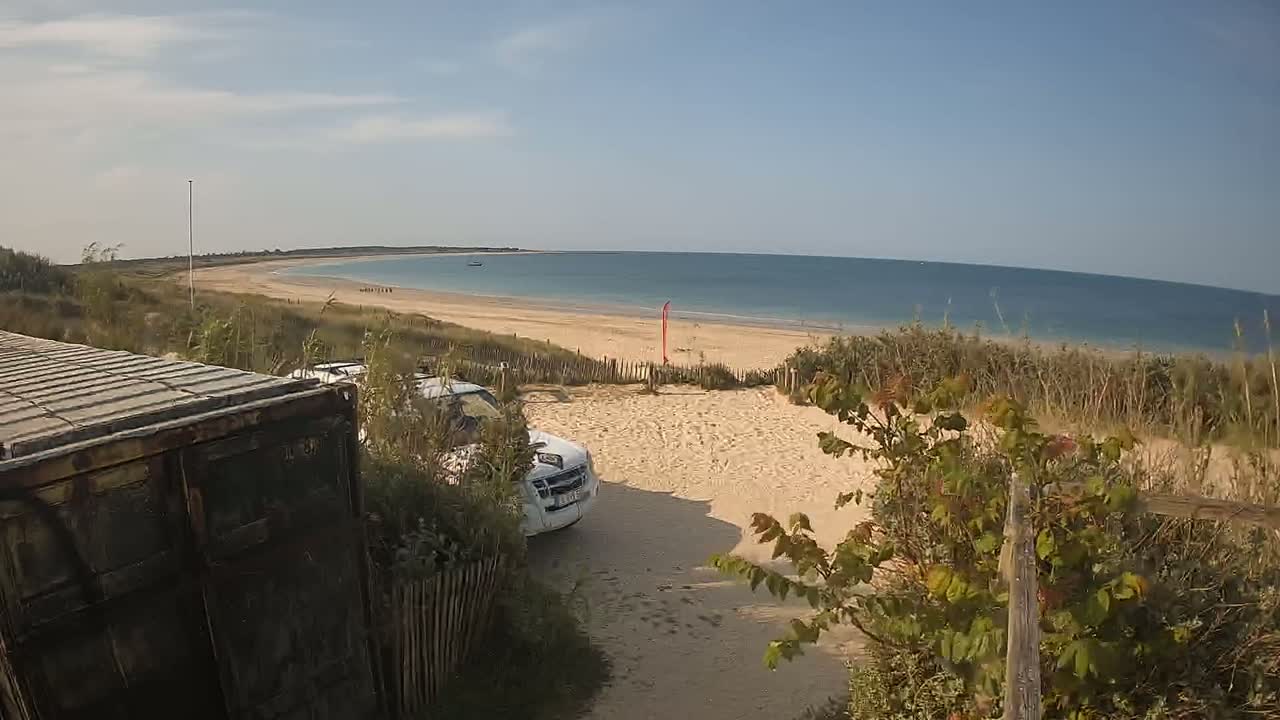 A sandy beach with calm blue water is visible beyond a dune, where a white vehicle is parked near a dark-colored shipping container; the weather is sunny.
