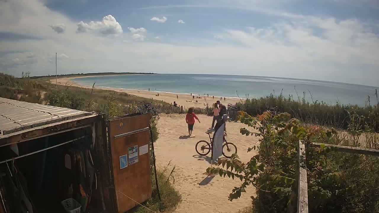 A sandy path leads to a beach with people swimming and sunbathing under a partly cloudy sky.