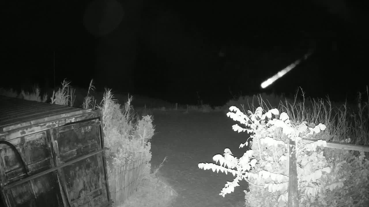 A bright meteor streaks across a dark night sky above a metal container and some shrubs.