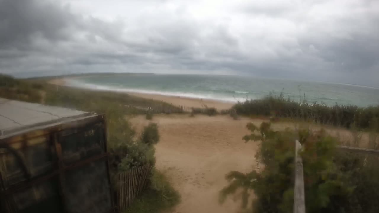 A sandy path leads to a beach with wavy ocean under a cloudy, overcast sky.
