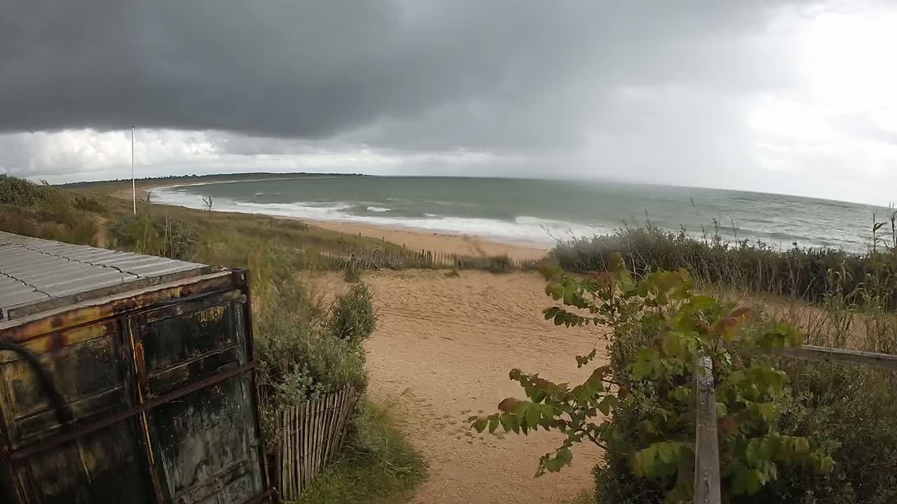 A weathered container sits beside a sandy path leading to a beach with choppy waves under a dark, overcast sky.