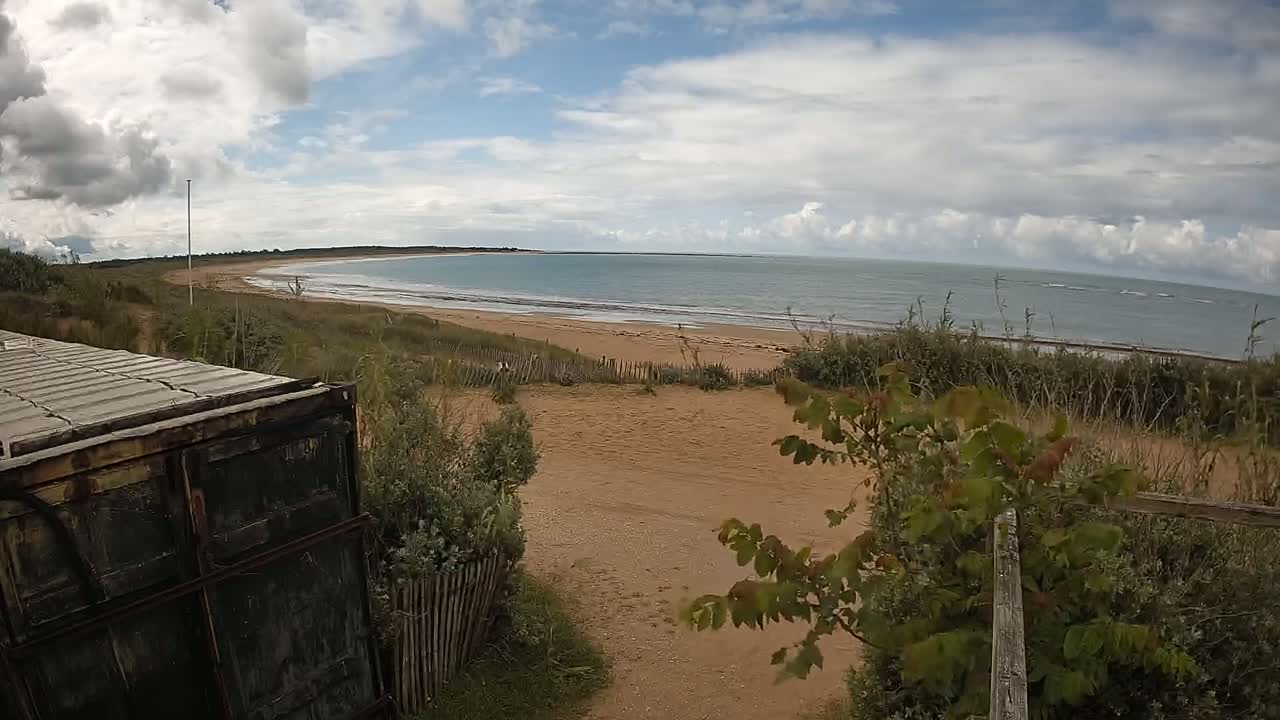A partly cloudy day reveals a view of a sandy beach and calm ocean, seen from behind a weathered dark container partially obscuring the foreground.
