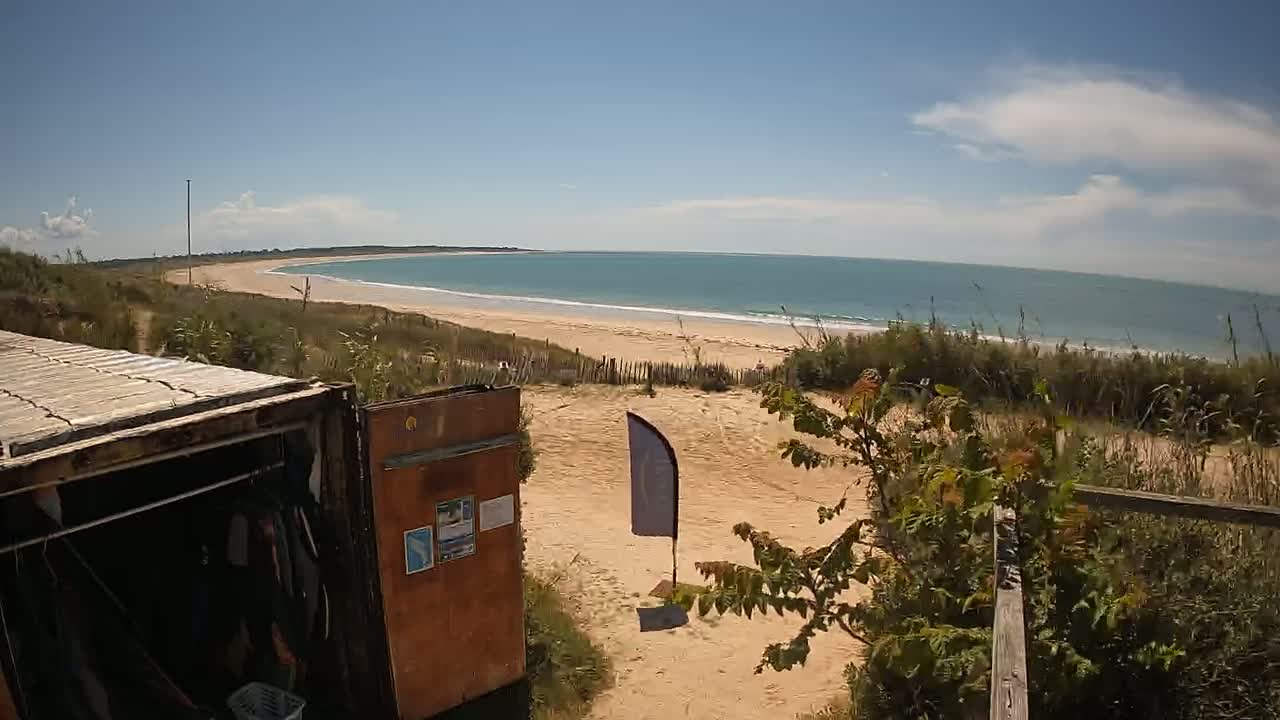 A sandy beach with a calm ocean stretches out under a partly cloudy sky, viewed from behind a weathered wooden container and dune vegetation.