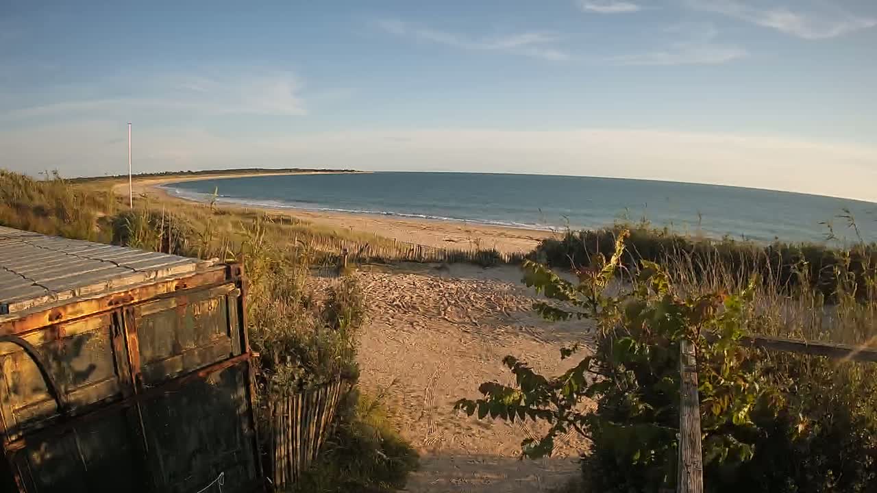 An aged metal container sits near a sandy path leading to a calm ocean beach under a mostly clear sky.