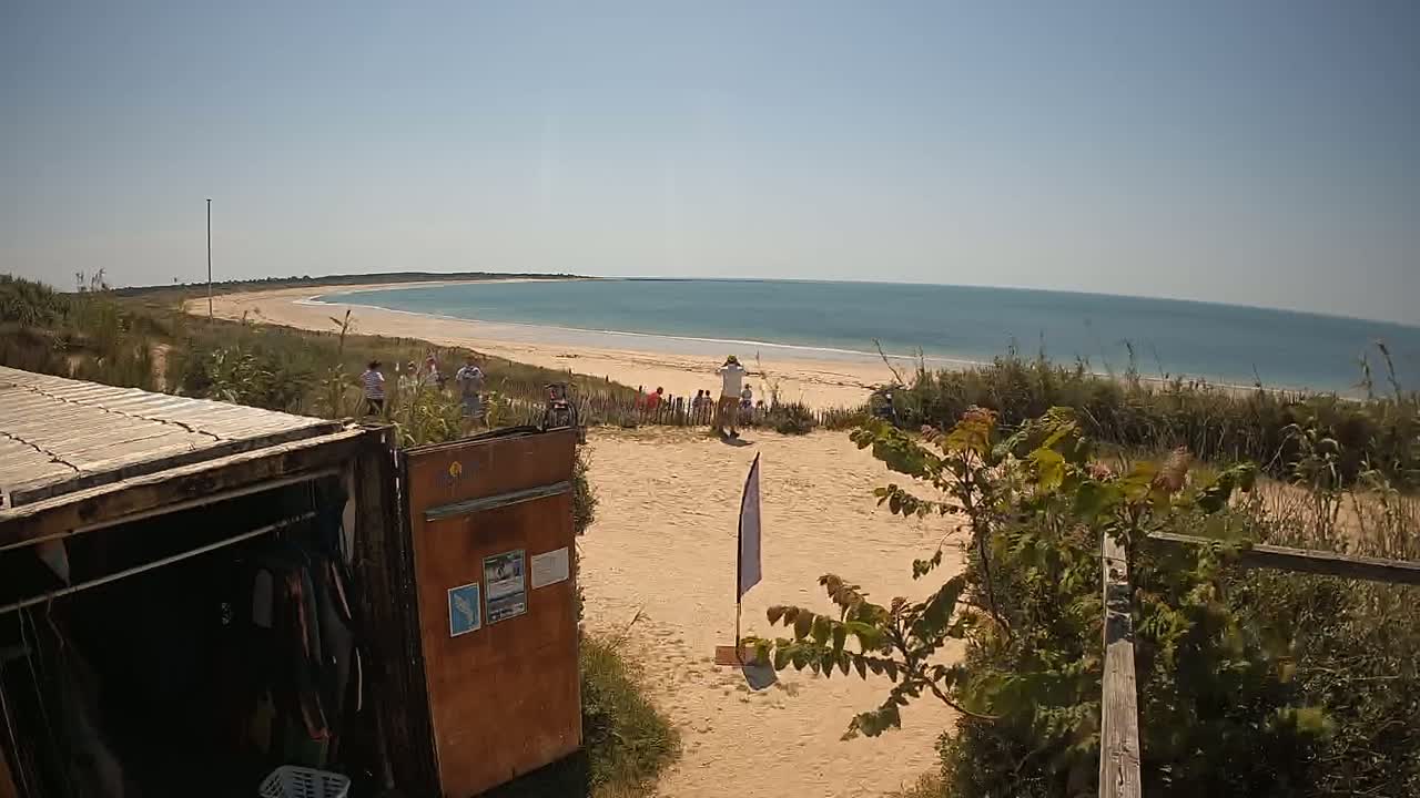 A sandy area overlooks a calm ocean beach on a sunny day, with several people gathered near the shoreline and a weathered storage container in the foreground.