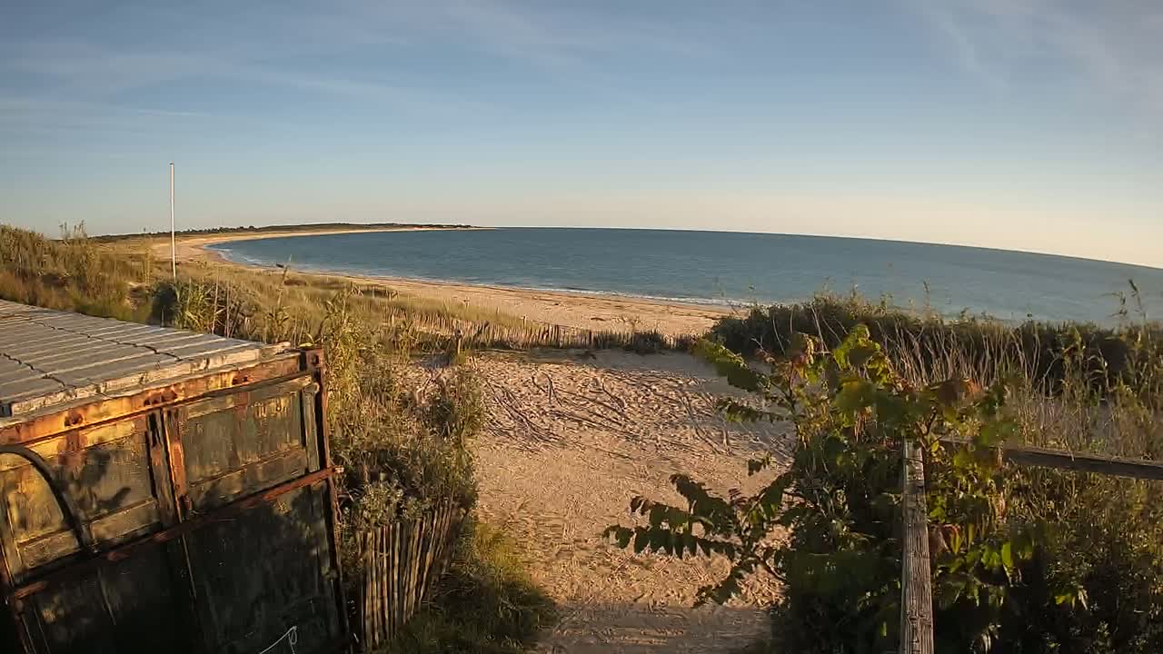 A rusty container sits near a sandy path leading to a calm ocean beach on a sunny day.