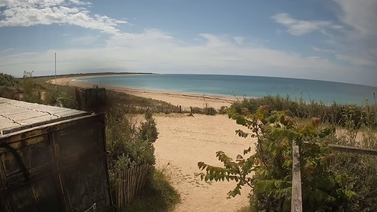 A sandy beach with calm, light-blue water stretches to a distant shoreline under a partly cloudy sky, viewed from behind a weathered, dark-colored container and low vegetation.