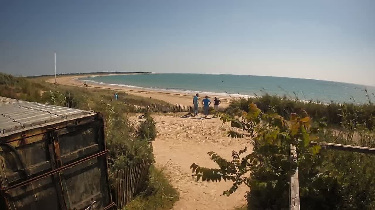 A sandy path leads to a calm, blue ocean beach where a few people stand near the shoreline on a sunny day.