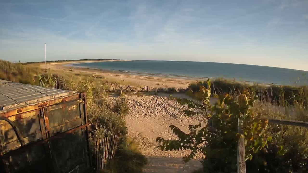 A rusty container sits near a sandy path leading to a calm, light-blue ocean beach on a sunny day.