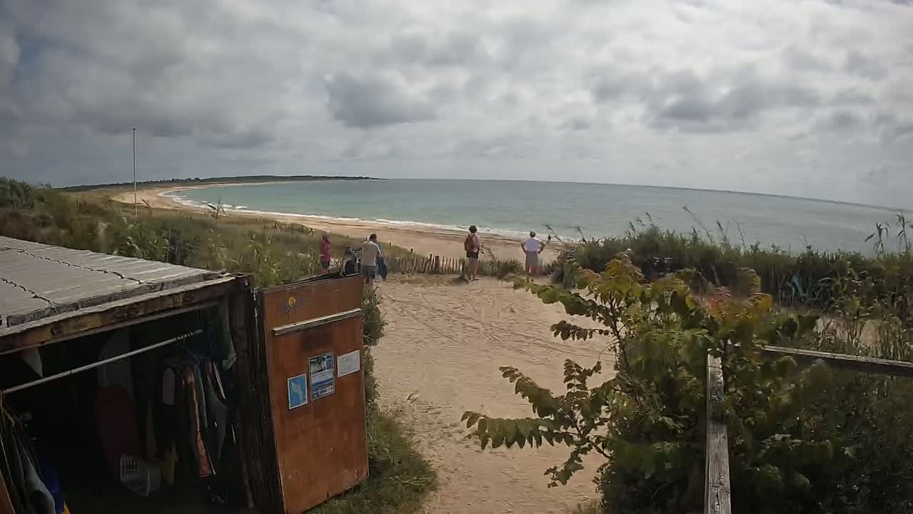 A sandy beach with calm ocean waves under a cloudy sky is viewed from behind a weathered shipping container with clothes hanging inside, near several people standing on a dune.