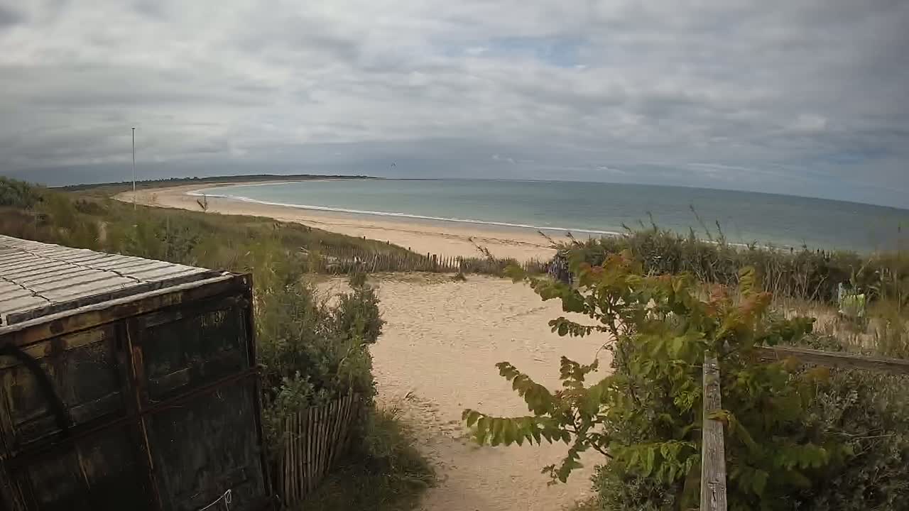 A view from behind a dark container shows a sandy beach and calm ocean under a cloudy sky.