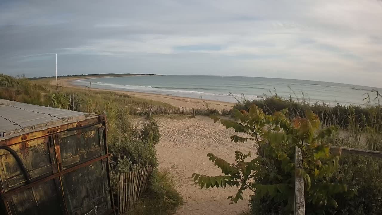 A view from behind a rusty container shows a sandy beach with gentle waves under a mostly cloudy sky.