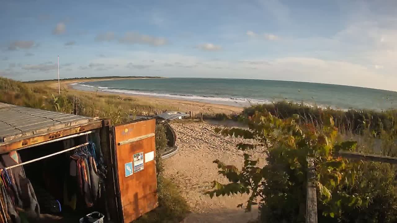A sandy beach with calm ocean waves is visible beyond a shipping container modified into a changing room, parked near a vehicle.