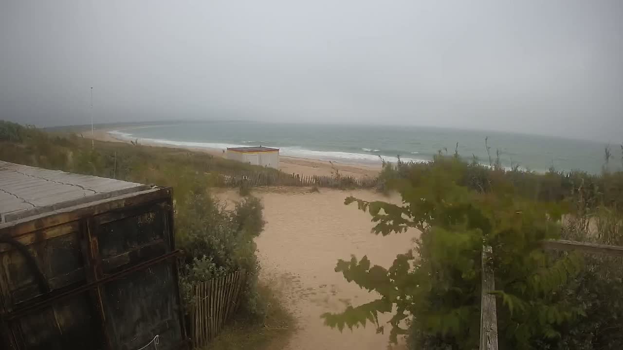 A view of a sandy beach with a small building and some vegetation, overlooking a wavy ocean under an overcast sky.