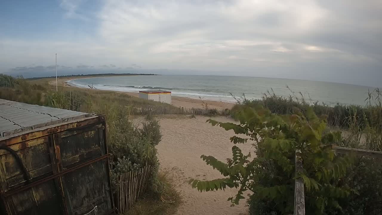 A sandy beach with a small building and a calm, overcast ocean is viewed from behind a rusty container.