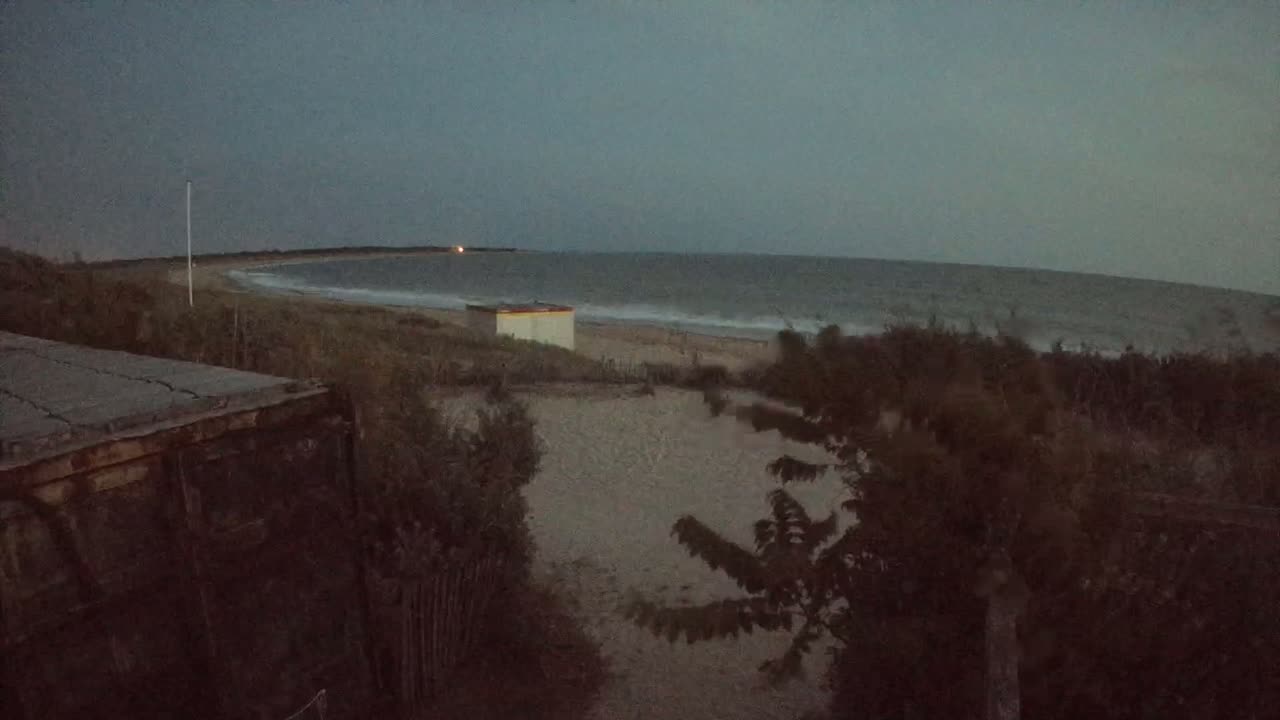 A dark, overcast beach scene shows a sandy shore, a small building, and a gray, wavy ocean under a gloomy sky.