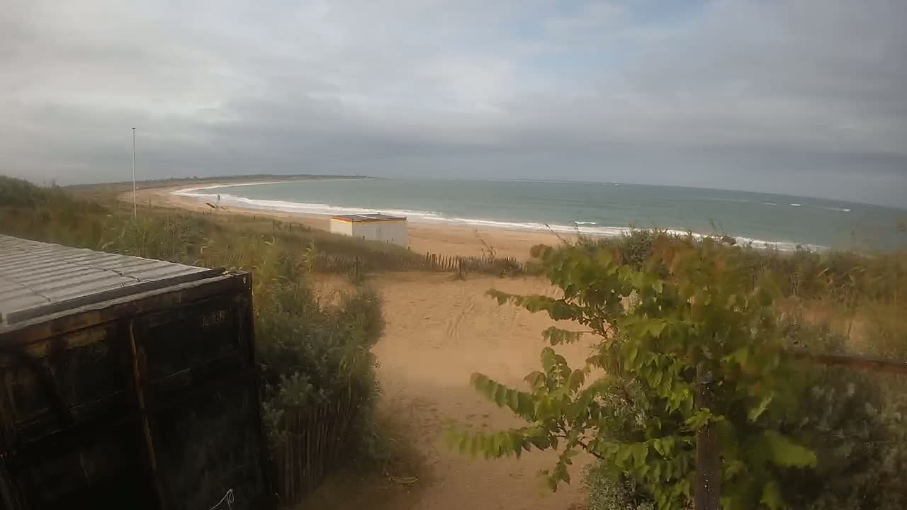 A sandy beach with a small building and a calm sea under an overcast sky is viewed from behind a dark container, partially obscured by vegetation.