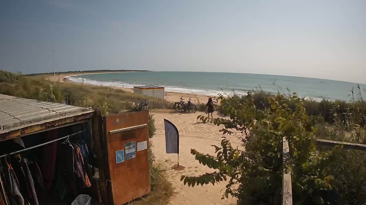 A sandy beach with a calm ocean stretches to a curved horizon under a clear sky, with a small building, bicycles, and a person visible near a repurposed shipping container used as a storage unit.