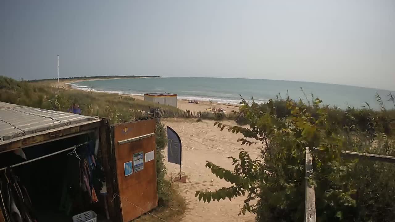 A sandy beach with calm ocean waves is visible beyond a path leading to a small building and a shipping container used as a changing room, partially obscured by vegetation.