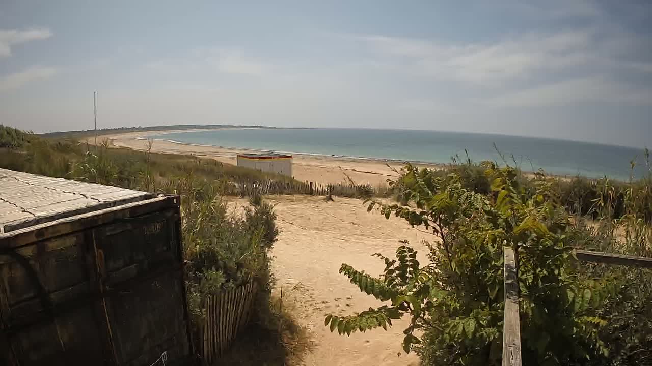 A sandy path leads to a beach with calm, light-blue water under a mostly sunny sky; a dark-colored container is partially visible in the foreground.