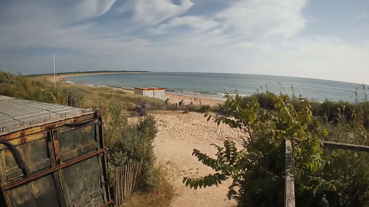 A rusty shipping container sits near a sandy path leading to a beach with calm ocean waves under a partly cloudy sky.