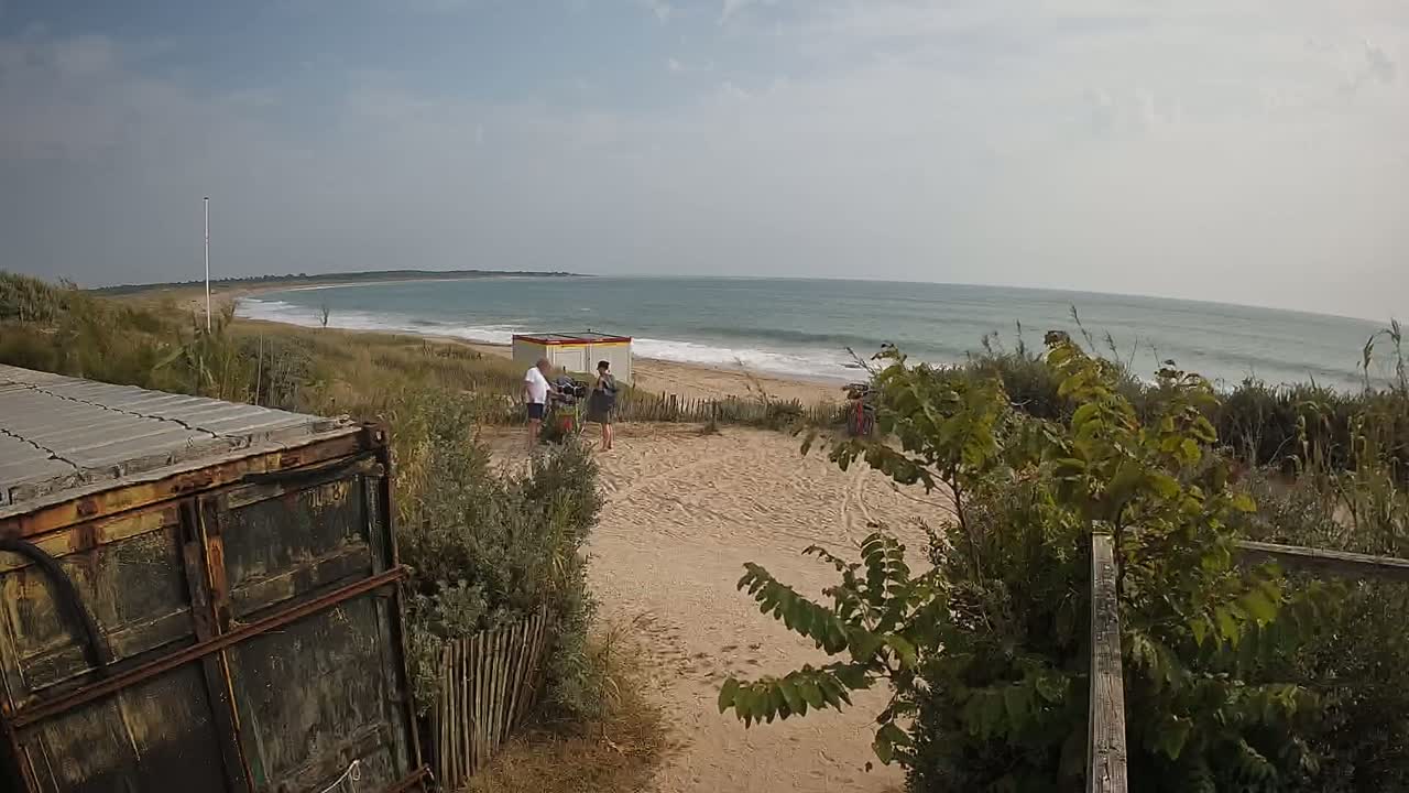 A sandy beach with small waves under a partly cloudy sky is seen from behind a rusty container, with two people standing near a small building in the middle ground.