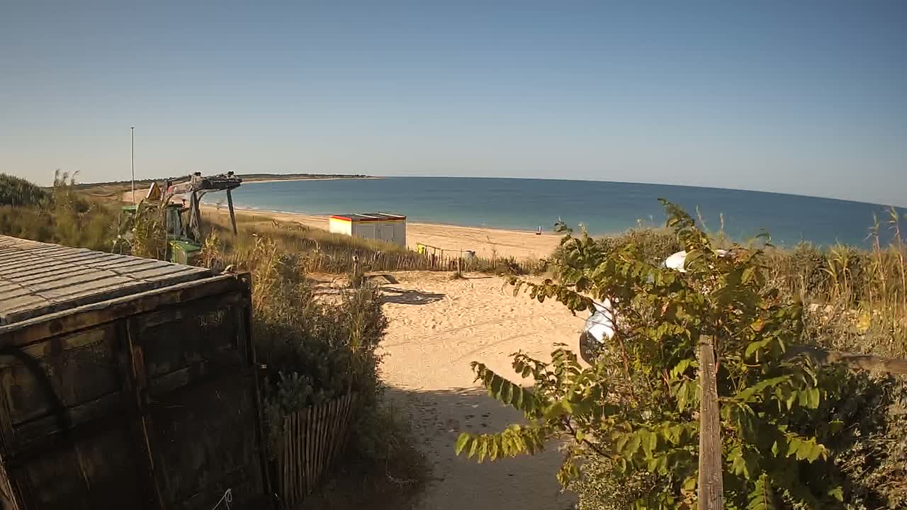 A sandy beach with calm, blue water is visible beyond a small dune, where construction equipment and a small building are situated near some vegetation and a parked vehicle.