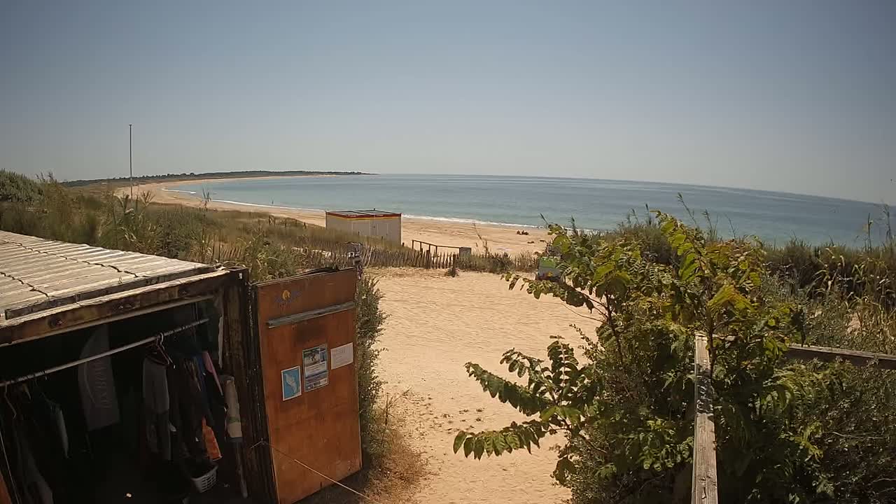 A sandy beach with calm ocean waves is seen from behind a weathered shipping container used as a changing room, on a sunny day.