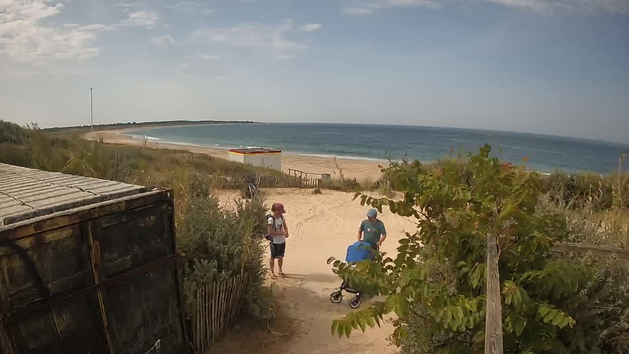 Two people stand near a sandy path leading to a beach with a calm ocean under a partly cloudy sky, while a third person pushes a stroller nearby.