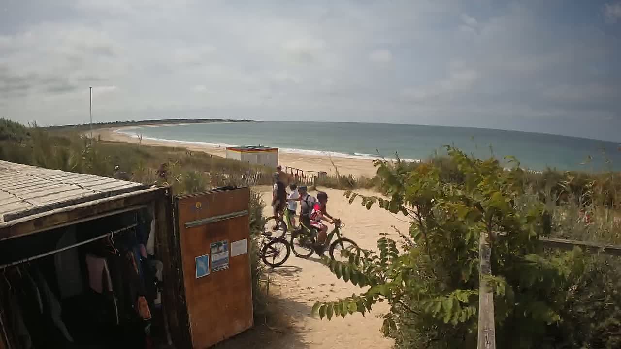 A group of people on bicycles ride along a sandy path toward a beach on a partly cloudy day.
