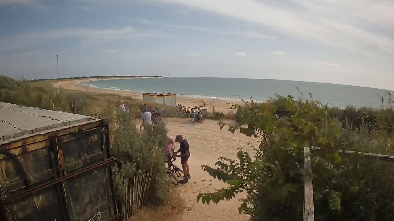 A sandy beach with calm ocean water under a partly cloudy sky is viewed from behind vegetation and a rusty shipping container, with several people near bicycles and a small building.