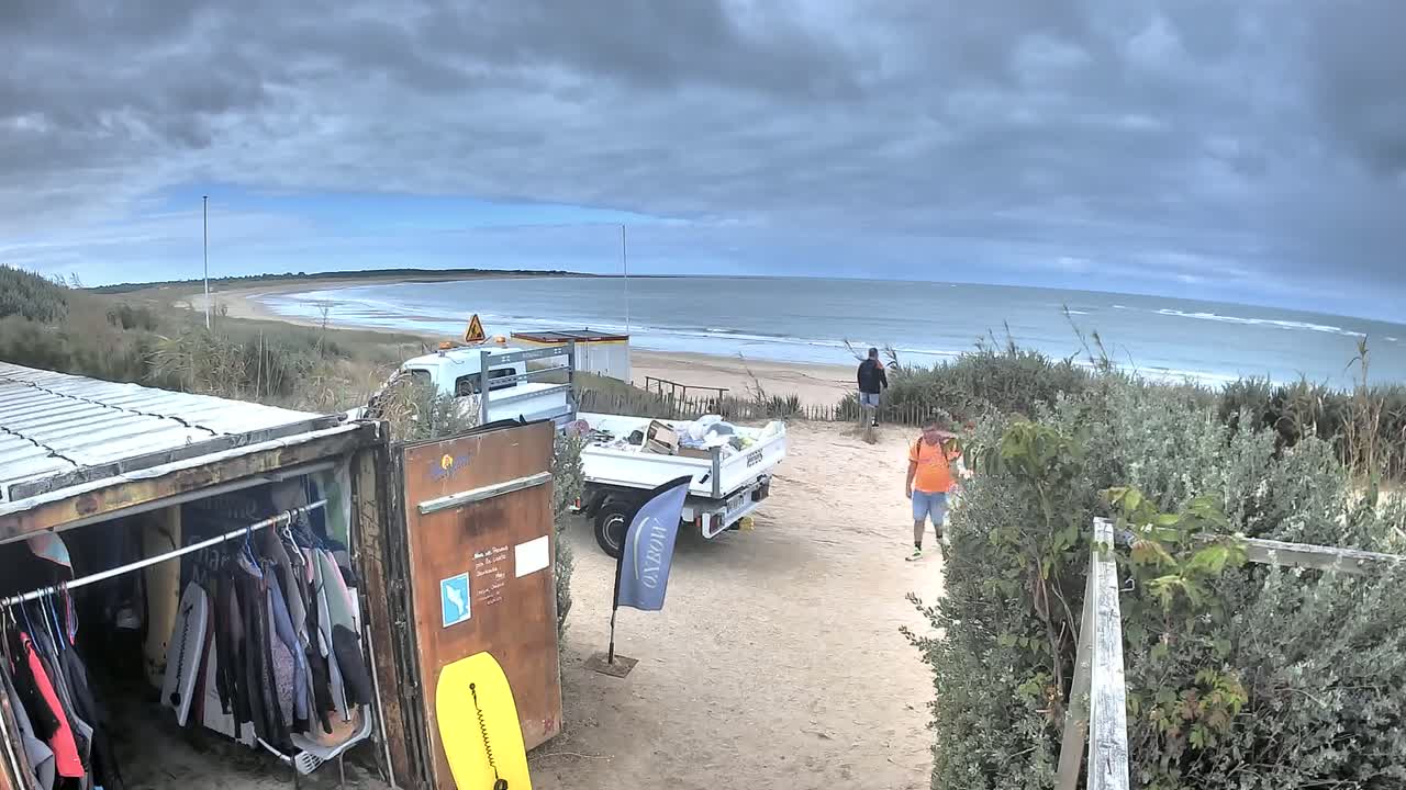 A sandy beach with an overcast sky is visible beyond a shipping container modified as a changing room, a parked truck, and several people near some vegetation.