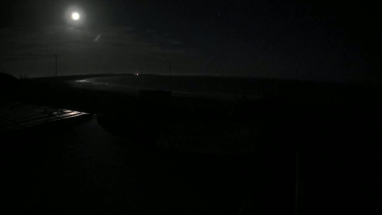 A bright moon illuminates a dark, nighttime scene of a body of water near a landmass under a partly cloudy sky.