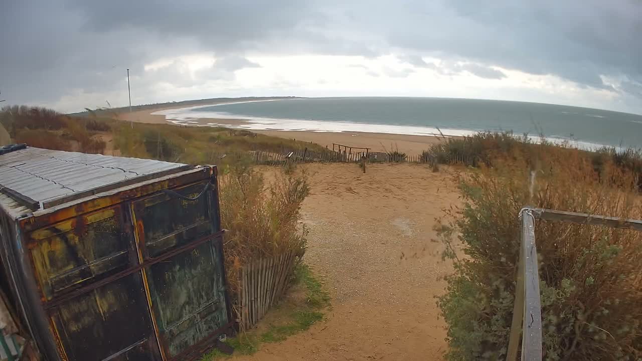 A wide, curving sandy beach and ocean stretch under a heavily overcast sky, viewed from a sandy dune with a rusty corrugated metal structure, scrubby vegetation, and a wooden fence in the foreground.