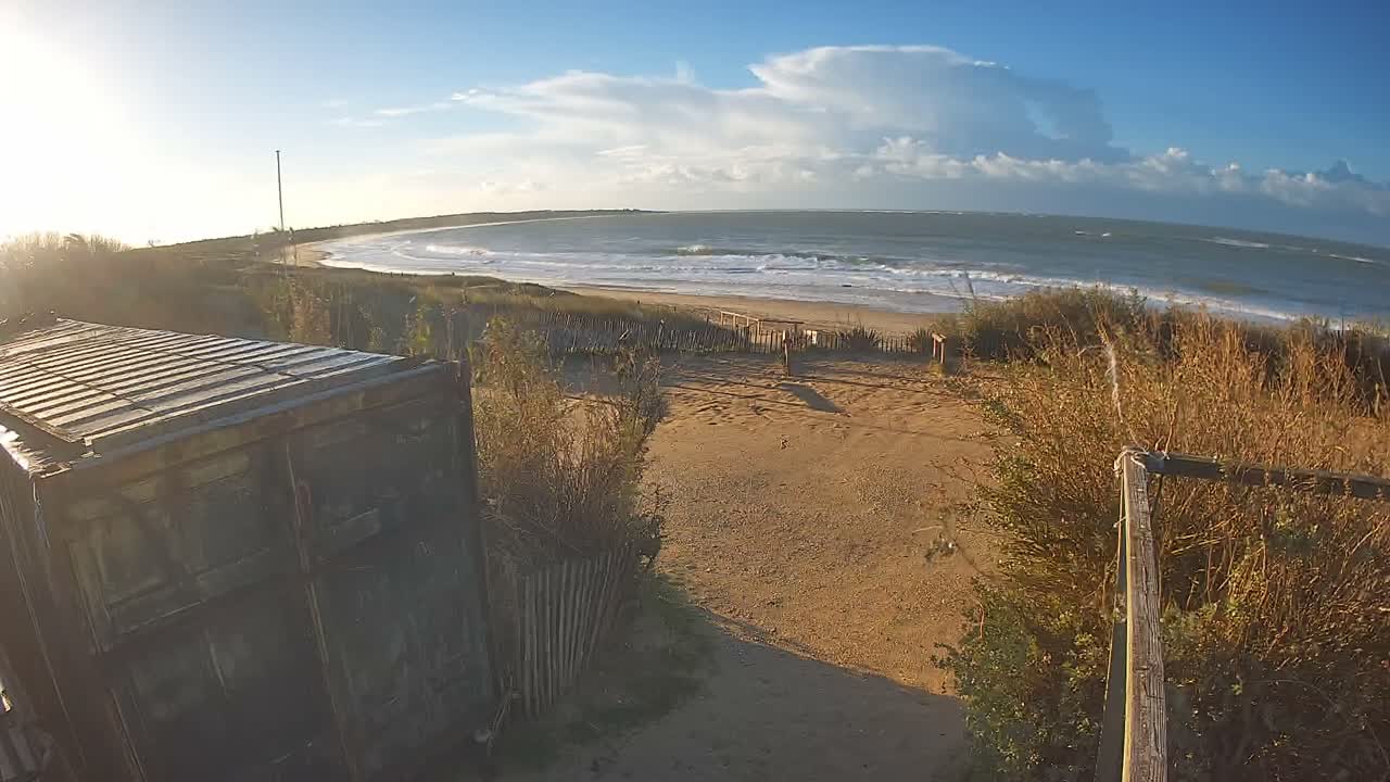 A clear and sunny day with scattered clouds illuminates a wide, curved sandy beach with crashing waves, bordered by vegetated dunes featuring a path and wooden fencing, with a weathered corrugated metal structure in the foreground.