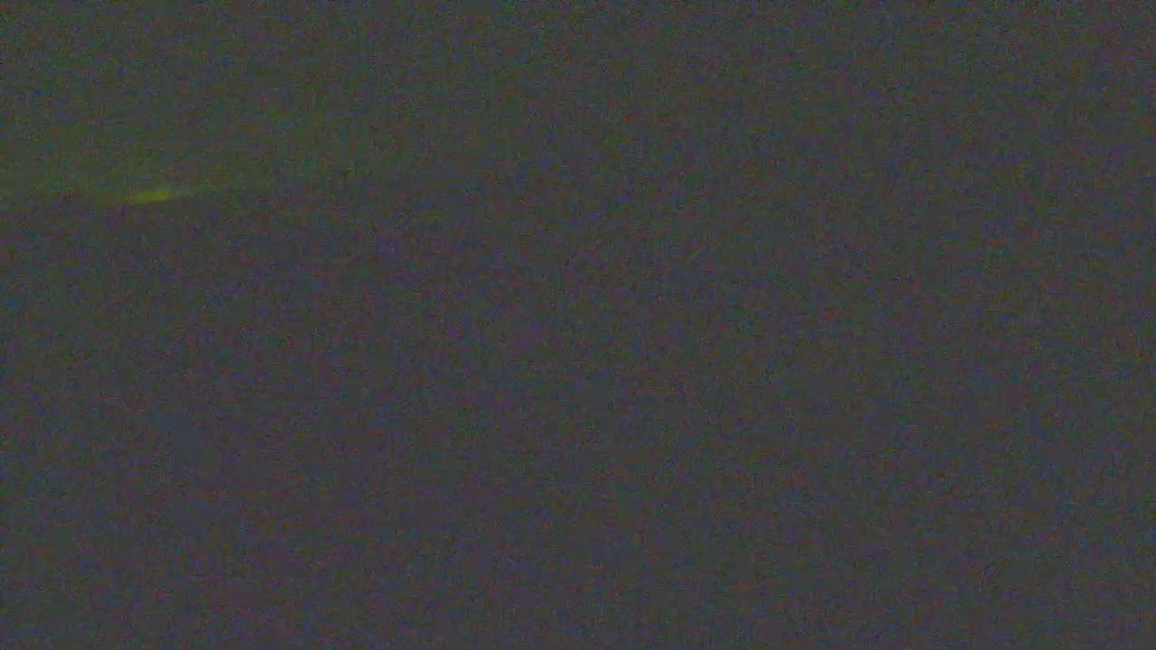 A clear and sunny day with scattered clouds illuminates a wide, curved sandy beach with crashing waves, bordered by vegetated dunes featuring a path and wooden fencing, with a weathered corrugated metal structure in the foreground.