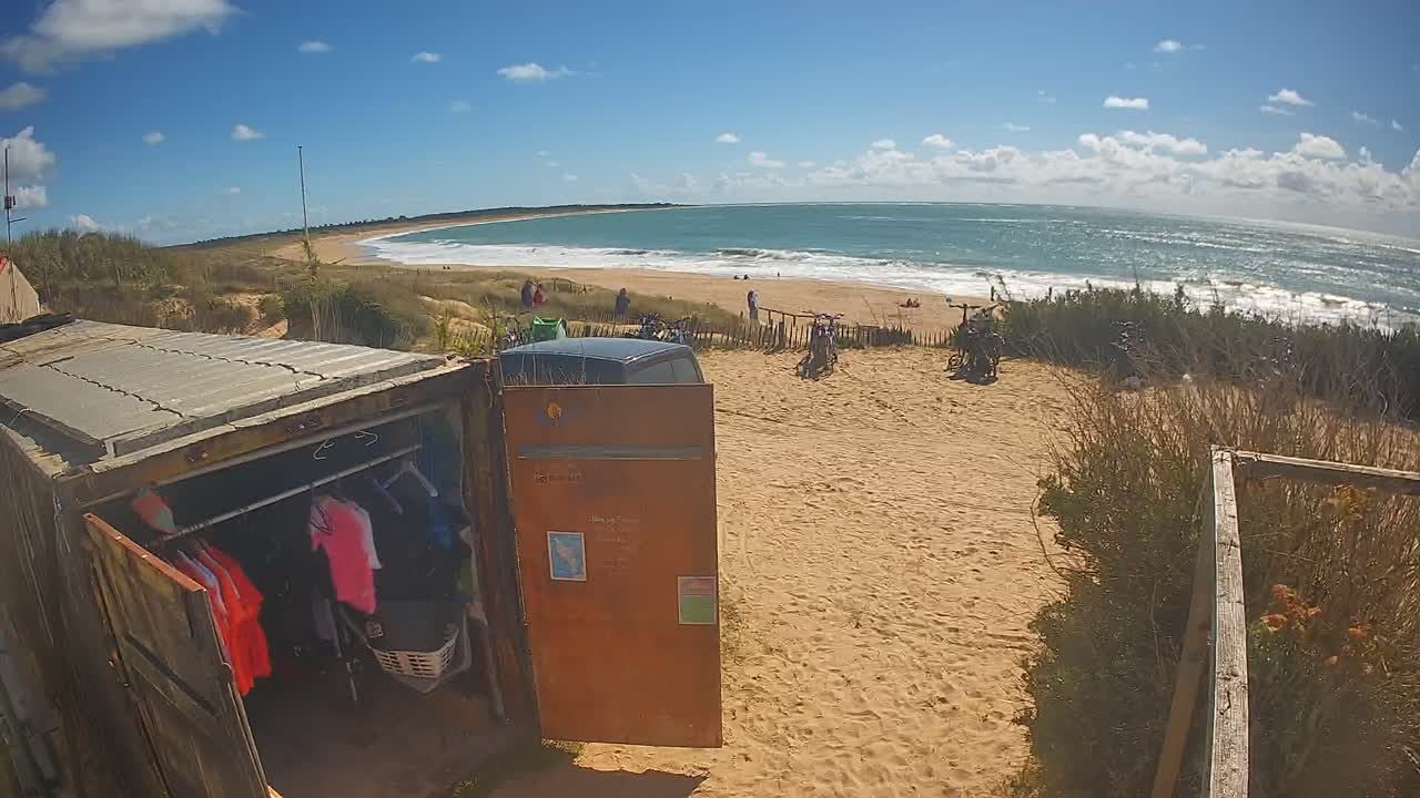 A clear and sunny day with scattered clouds illuminates a wide, curved sandy beach with crashing waves, bordered by vegetated dunes featuring a path and wooden fencing, with a weathered corrugated metal structure in the foreground.
