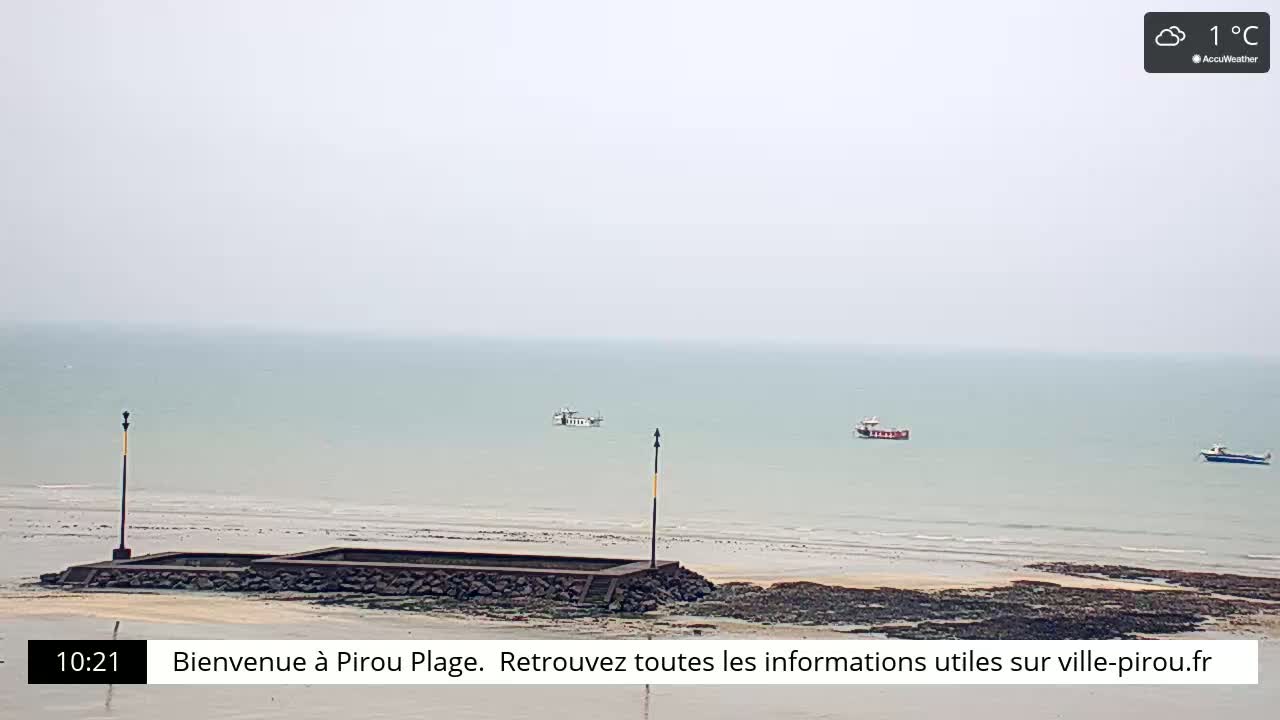 A hazy, overcast day at a beach shows calm, pale-green water with a few small boats and a low stone wall on the shore.